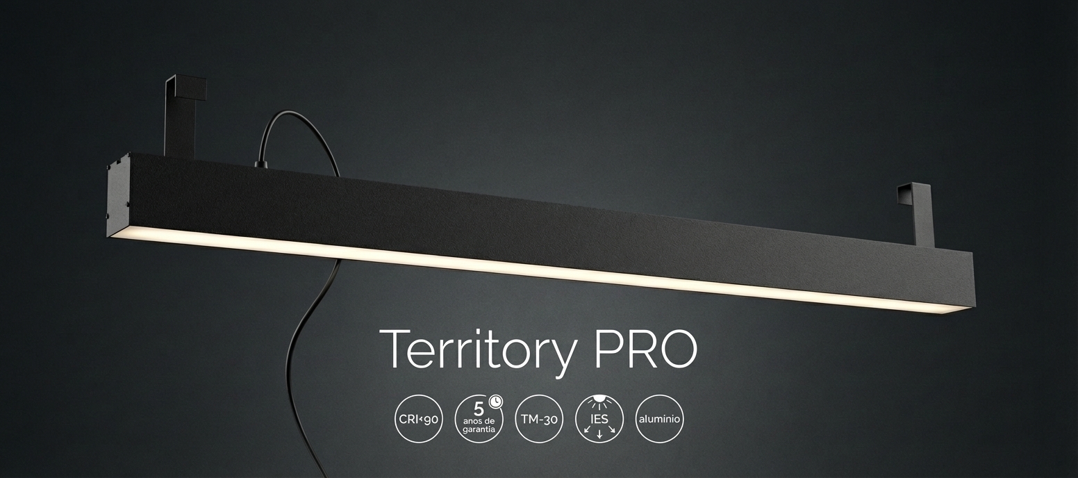 Imagem promocional de uma luminária pendente linear preta, com design moderno e minimalista, suspensa por suportes rígidos e cabo elétrico aparente. A peça apresenta iluminação LED difusa na parte inferior, destacando seu acabamento sofisticado em alumínio. Ao fundo escuro, o nome “Territory PRO” aparece em destaque, acompanhado de ícones com especificações como CRI>90, 5 anos de garantia, TM-30, IES e material em alumínio.